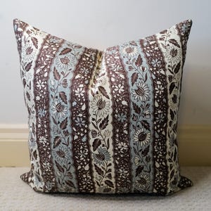 May include: A decorative pillow with a floral pattern. The pillow features vertical stripes of brown, light blue, and cream colors. The floral design includes stylized flowers and leaves, creating a vintage aesthetic. The pillow is square-shaped and appears to be made of fabric.