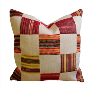 May include: Square cushion with a patchwork design. The cushion has a cream-coloured background with squares of red, yellow, and brown stripes. The cushion is square and appears to be made of a woven fabric.
