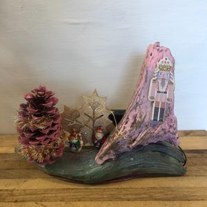 May include: A decorative Christmas display featuring a pink painted wooden base with a pine cone, a leaf, and miniature figurines. A pink painted wooden piece with a nutcracker is on the right. Gold string lights are wrapped around the display.