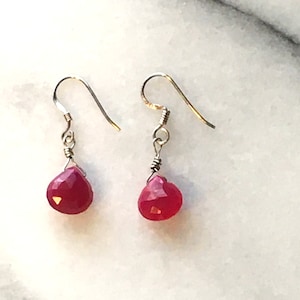 May include: A pair of drop earrings featuring faceted, teardrop-shaped, red gemstones. The earrings have silver-colored ear wires and are displayed against a white and gray marble background. The gemstones are approximately 0.5 inches in length.