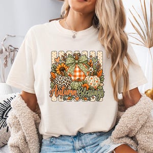 May include: A cream-colored t-shirt featuring a graphic print of three pumpkins with varied patterns, including leopard print and orange plaid. The design includes a large green polka-dot bow, sunflowers, leaves, and the text Autumn Blessings.