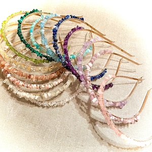 Può includere: Una collezione di cerchietti per capelli con pietre preziose in vari colori, tra cui verde, blu, viola e rosa. Ogni cerchietto presenta una sottile fascia metallica dorata adornata con piccole pietre di forma irregolare. I cerchietti sono disposti in una mostra a strati.