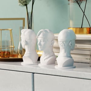 May include: Three white ceramic busts of people wearing military hats. The busts are displayed on a white shelf.