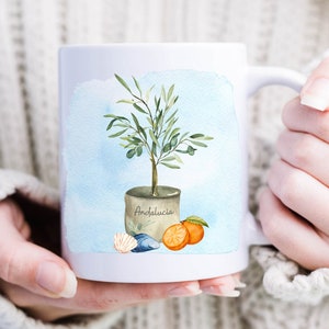 May include: White ceramic mug with a watercolor illustration of an olive tree in a pot with the word "Andalucia" on the pot. The tree is surrounded by oranges and seashells.