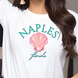 Puede incluir: Camiseta blanca con la palabra "NAPLES" en verde azulado sobre un gráfico de concha rosa. Debajo, la palabra "Florida" está escrita en cursiva verde azulado. La camiseta se combina con vaqueros azul claro.