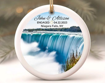 May include: A white ceramic ornament with a gold string for hanging. The ornament features a scenic image of Niagara Falls with the text "John & Allison ENGAGED 04.22.2023 Niagara Falls, NY".