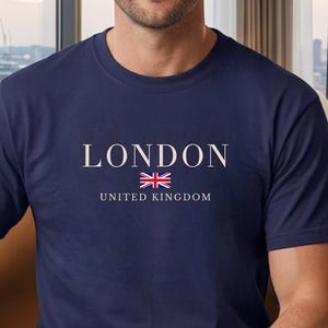以下が含まれることがあります： ネイビーブルーの半袖Tシャツ。胸の中央に明るい色のフォントで「LONDON」という文字、小さなユニオンジャックの旗のグラフィック、そしてそれより小さい明るい色のフォントで「UNITED KINGDOM」という文字がプリントされています。