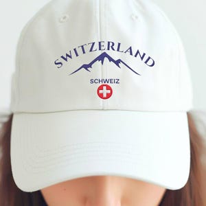 May include: White baseball cap with the word "SWITZERLAND" arched across the top in navy blue, with a mountain range graphic. Below the word "SWITZERLAND" is the word "SCHWEIZ" and a red and white cross emblem.