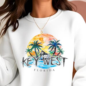 May include: White sweatshirt featuring a colorful Key West, Florida design. The graphic includes a sunset, palm trees, a small house, and the text "KEY WEST FLORIDA" in a stylized font. The sweatshirt is a casual, comfortable style.