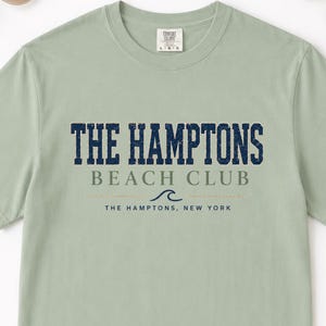 May include: A sage green short-sleeve t-shirt featuring dark blue distressed text that reads THE HAMPTONS BEACH CLUB, with a small wave graphic and the text THE HAMPTONS, NEW YORK printed below in a smaller, thinner font on the chest area.