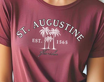 Może przedstawiać: Bordowa koszulka z podwiniętymi rękawami z napisem "ST. AUGUSTINE" w zakrzywionej białej czcionce, grafiką palm i napisem "EST. 1565 Florida". Koszulka wykonana jest z miękkiego materiału.
