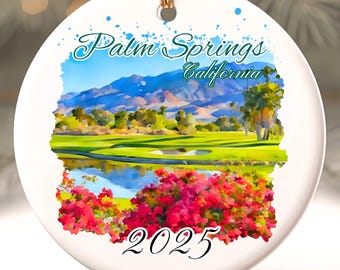 Op de afbeelding: Een ronde witte keramische kerstversiering met een aquarel schilderij van Palm Springs, Californië. De afbeelding toont bergen, een golfbaan en rode bloemen. De tekst "Palm Springs California" en "2025" zijn zichtbaar.