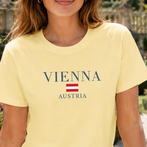 Könnte beinhalten: Ein hellgelbes Kurzarm-T-Shirt mit dem Text VIENNA in dunkelblauen Großbuchstaben, einem kleinen horizontalen rot-weißen österreichischen Flaggensymbol und dem Wort AUSTRIA in kleineren dunkelblauen Großbuchstaben mittig auf der Brust.