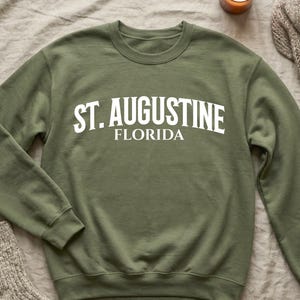 May include: Military green sweatshirt with the text "ST. AUGUSTINE FLORIDA" in white. The sweatshirt is on a neutral-colored surface, with a lit candle, a folded sweater, and a pair of socks.
