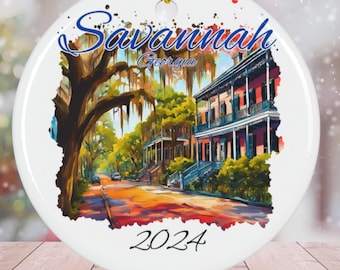 Puede incluir: Adorno navideño redondo blanco con una pintura de acuarela de una escena callejera en Savannah, Georgia. La pintura presenta un árbol con musgo español, una calle de adoquines y edificios históricos. El texto "Savannah Georgia" y "2024" está impreso en el adorno.