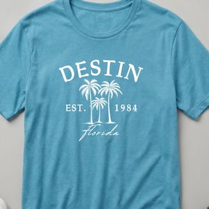 May include: A turquoise t-shirt featuring the word "DESTIN" in white, with "EST. 1984" and "Florida" below a palm tree graphic. The shirt is displayed on a neutral background with a watch, belt, and shoes.