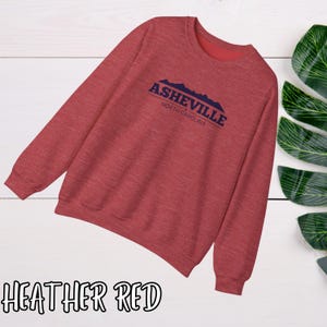 K&ouml;nnte beinhalten: Heather-roter Pullover mit einem blauen Grafikdruck, auf dem "Asheville, North Carolina" mit einer Bergsilhouette steht.
