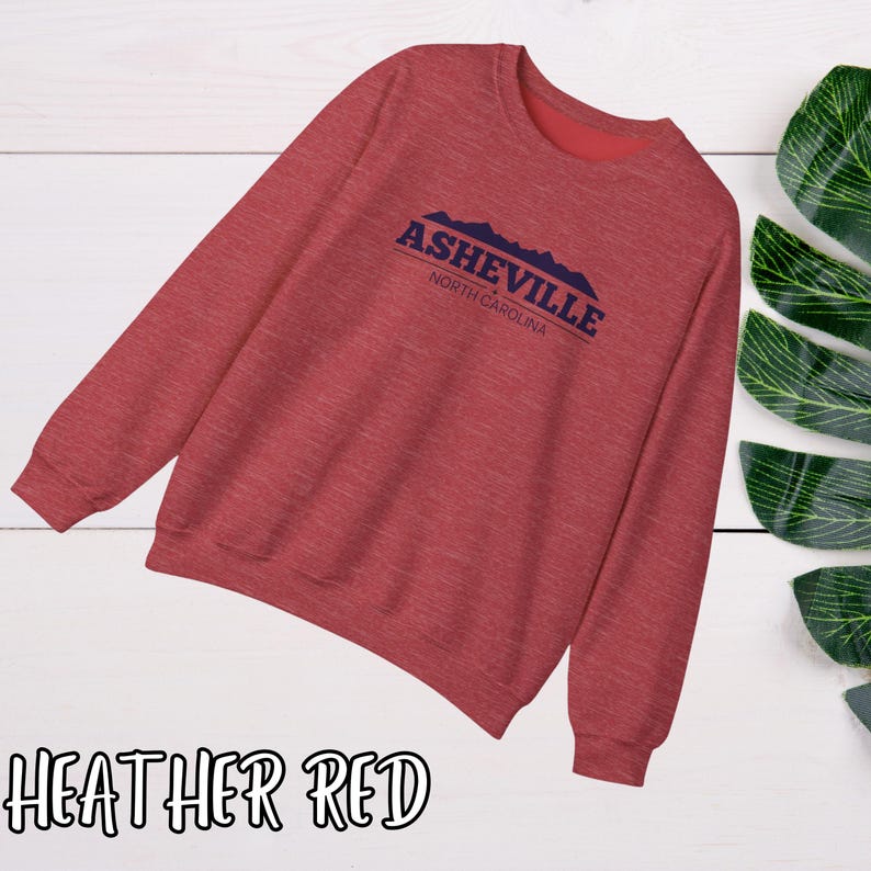 K&ouml;nnte beinhalten: Heather-roter Pullover mit einem blauen Grafikdruck, auf dem "Asheville, North Carolina" mit einer Bergsilhouette steht.
