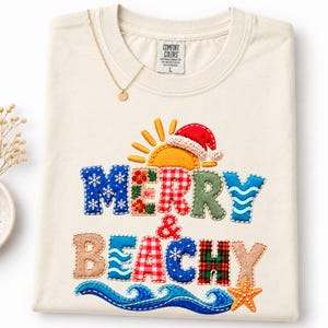 May include: A cream-coloured t-shirt featuring a graphic with the text Merry & Beachy. The design includes a yellow sun wearing a red Santa hat, blue snowflake patterns, red and white gingham, green textures, and a blue ocean wave with a starfish.