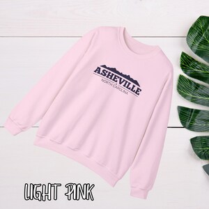 K&ouml;nnte beinhalten: Hellrosafarbener Pullover mit einem marineblauen Aufdruck, der "Asheville, North Carolina" mit einer Bergsilhouette zeigt.