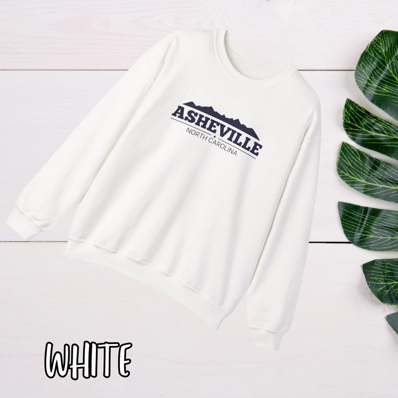 K&ouml;nnte beinhalten: Wei&szlig;er Pullover mit einem marineblauen Grafikdruck, der "Asheville, North Carolina" mit einer Silhouette von Bergen sagt.