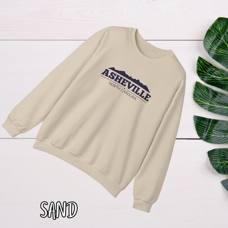 K&ouml;nnte beinhalten: Ein beiger Pullover mit einem marineblauen Grafikdruck, der "Asheville, North Carolina" mit einer Silhouette von Bergen sagt.