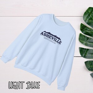 K&ouml;nnte beinhalten: Hellblauer Pullover mit dem Text "Asheville, North Carolina" und einer Grafik von Bergen.