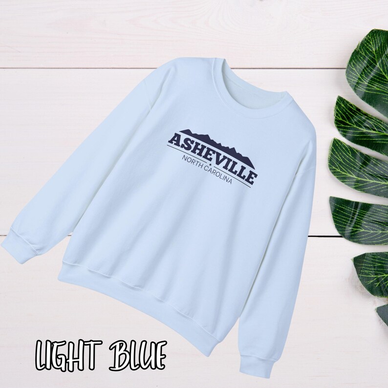 K&ouml;nnte beinhalten: Hellblauer Pullover mit dem Text "Asheville, North Carolina" und einer Grafik von Bergen.