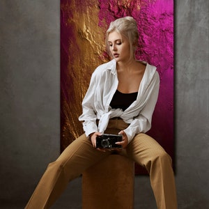 May include: A woman wearing a white shirt and brown pants sits on a wooden stool in front of a large abstract painting with gold and pink colors. She is holding a vintage camera in her hands.