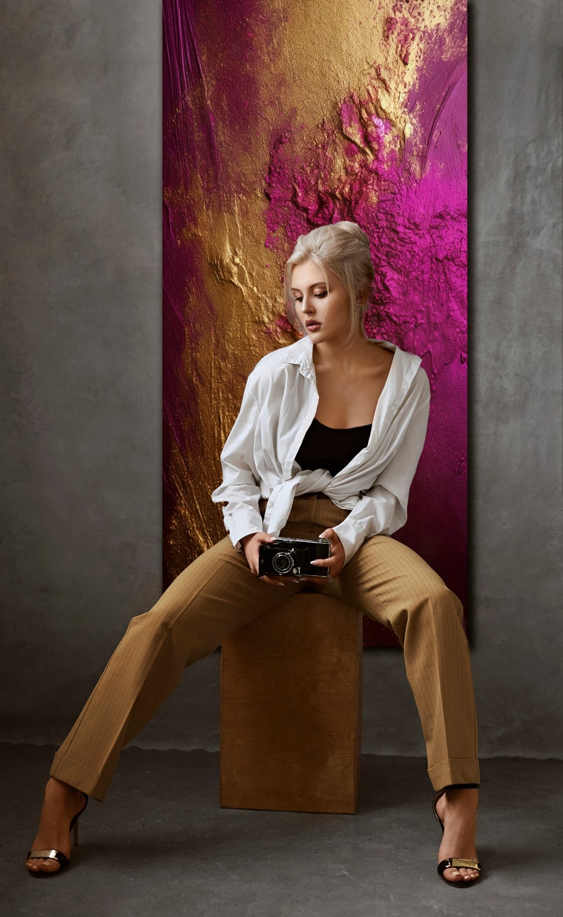 May include: A woman wearing a white shirt and brown pants sits on a wooden stool in front of a large abstract painting with gold and pink colors. She is holding a vintage camera in her hands.