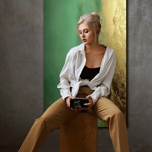 May include: A woman wearing a white shirt, brown pants, and black heels sits on a wooden stool. She is holding a vintage camera in front of her. The background is a green and gold abstract painting.