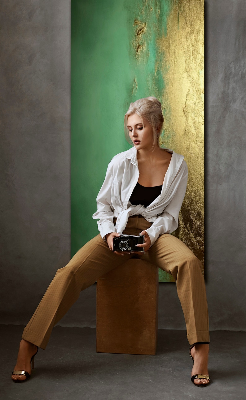 May include: A woman wearing a white shirt, brown pants, and black heels sits on a wooden stool. She is holding a vintage camera in front of her. The background is a green and gold abstract painting.