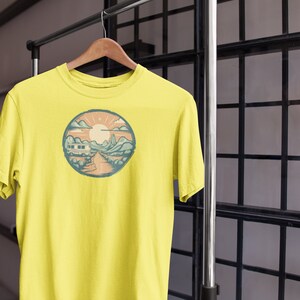 May include: A yellow t-shirt featuring a circular graphic of a mountain landscape with a camper van driving on a road. The sun is shining in the sky.