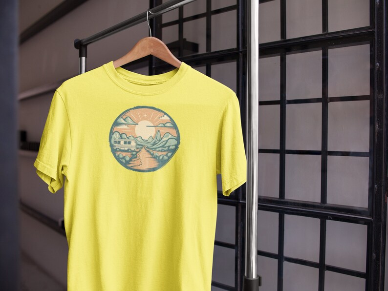 May include: A yellow t-shirt featuring a circular graphic of a mountain landscape with a camper van driving on a road. The sun is shining in the sky.