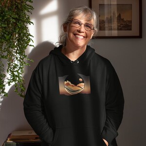 May include: A black hooded sweatshirt featuring a graphic print of a stylised landscape within a circle. The landscape is a blend of brown, orange, and blue colours.