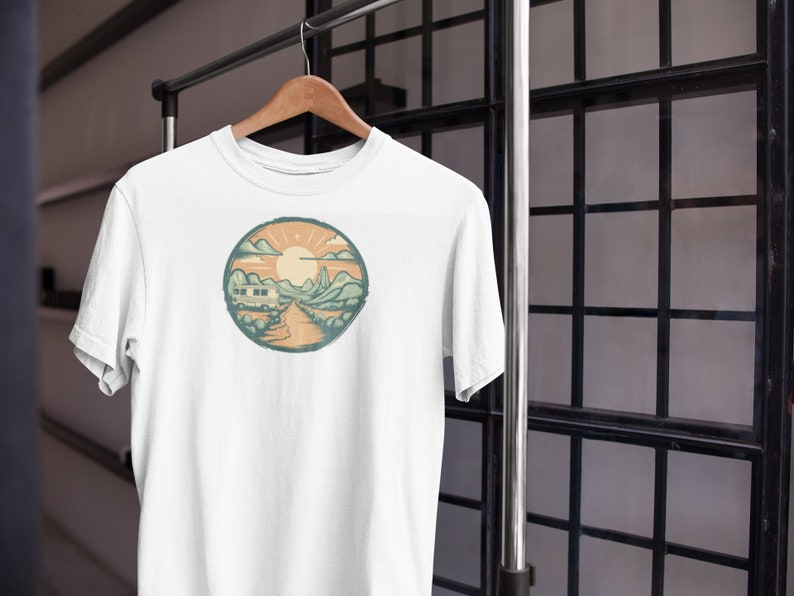 May include: White t-shirt featuring a circular graphic of a camper van driving through a mountain landscape with a sun and clouds in the background.