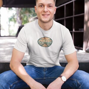 May include: A white t-shirt with a circular graphic design featuring a landscape with mountains, a sun, and a road. The graphic is in shades of green, brown, and orange.