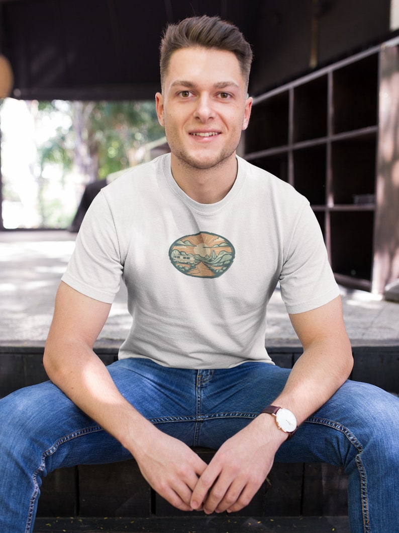 May include: A white t-shirt with a circular graphic design featuring a landscape with mountains, a sun, and a road. The graphic is in shades of green, brown, and orange.