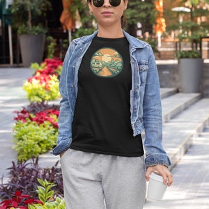 May include: A woman wearing a black t-shirt with a retro-style illustration of a camper van driving through a mountain landscape with a sun in the sky. The t-shirt has a round, orange border around the illustration. The woman is wearing a denim jacket and grey sweatpants. She is holding a white takeaway cup.
