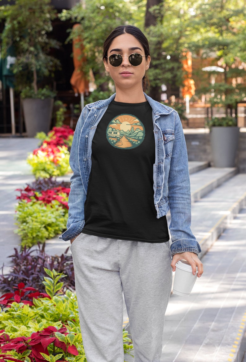 May include: A woman wearing a black t-shirt with a retro-style illustration of a camper van driving through a mountain landscape with a sun in the sky. The t-shirt has a round, orange border around the illustration. The woman is wearing a denim jacket and grey sweatpants. She is holding a white takeaway cup.