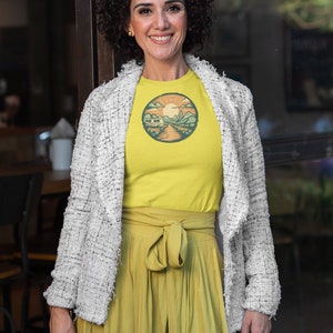 May include: A woman wearing a yellow t-shirt with a retro-style illustration of a camper van in a mountain landscape. The t-shirt has a round, orange-colored border. She is also wearing a white jacket and a yellow skirt.