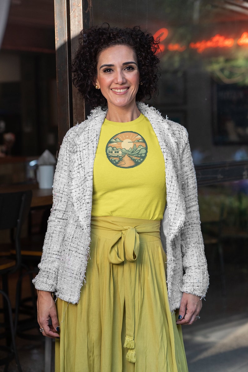 May include: A woman wearing a yellow t-shirt with a retro-style illustration of a camper van in a mountain landscape. The t-shirt has a round, orange-colored border. She is also wearing a white jacket and a yellow skirt.