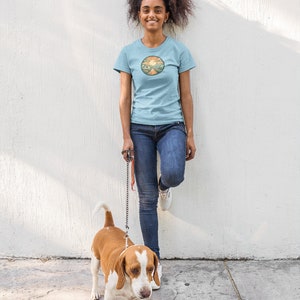 May include: A woman wearing a light blue t-shirt with a circular graphic of a sunset over a road. She is standing next to a white and brown beagle dog on a lead.
