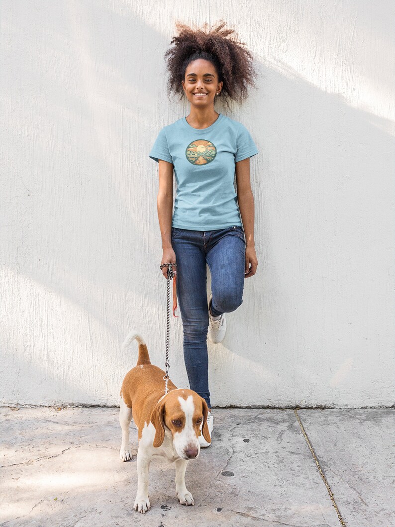 May include: A woman wearing a light blue t-shirt with a circular graphic of a sunset over a road. She is standing next to a white and brown beagle dog on a lead.