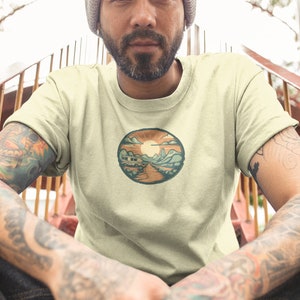May include: A light beige t-shirt featuring a circular graphic of a camper van driving down a road with mountains in the background. The sun is setting behind the mountains and there are clouds in the sky.