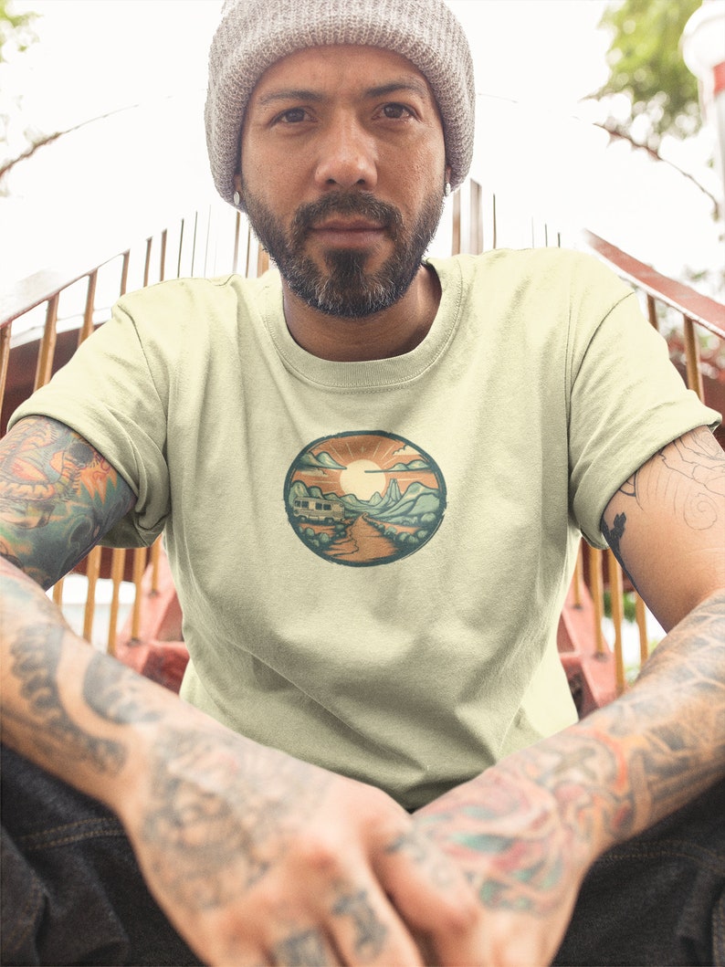 May include: A light beige t-shirt featuring a circular graphic of a camper van driving down a road with mountains in the background. The sun is setting behind the mountains and there are clouds in the sky.