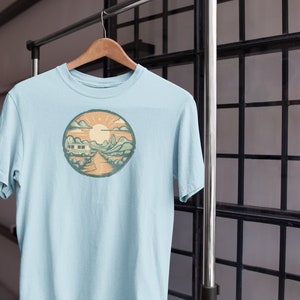 May include: A light blue t-shirt featuring a graphic print of a camper van driving through a mountain landscape with a sun and clouds in the background.