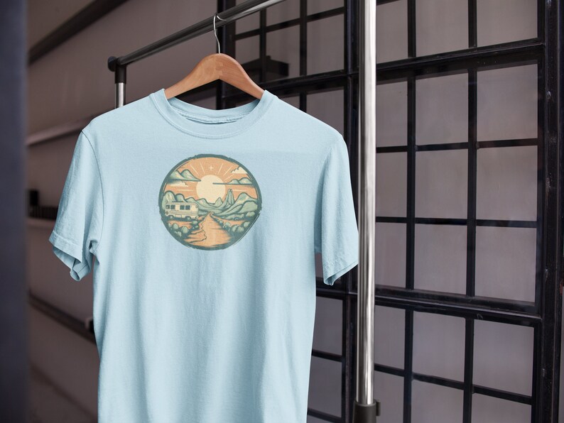 May include: A light blue t-shirt featuring a graphic print of a camper van driving through a mountain landscape with a sun and clouds in the background.