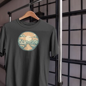 May include: A dark grey t-shirt featuring a vintage-style illustration of a camper van driving down a road through a mountain landscape with a sun in the sky.