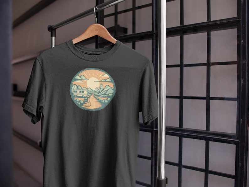 May include: A dark grey t-shirt featuring a vintage-style illustration of a camper van driving down a road through a mountain landscape with a sun in the sky.